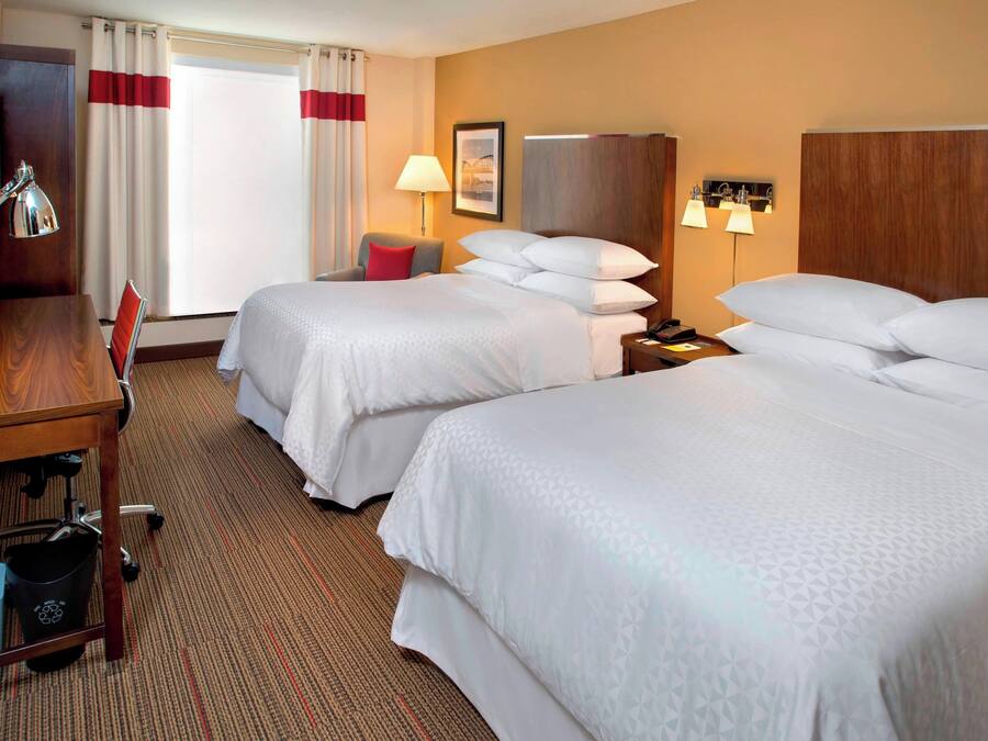 A hotel room with two beds, a desk, a chair, a TV, and a lamp.