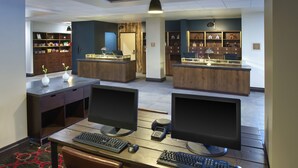Un espacio de oficina moderno con dos escritorios, cada uno con monitor, teclado y ratón. Hay dos sillas en cada escritorio. La habitación tiene estanterías de madera con varios objetos y una pequeña zona de recepción con un escritorio y una silla.