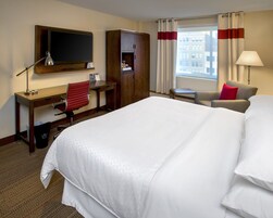 Habitación de hotel con una cama grande, un escritorio con silla, televisor, armario y ventana con cortinas.