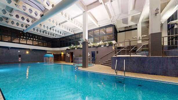 An indoor swimming pool with a clear view of the pool area, including the pool's edge, a set of stairs leading to a higher level, and a tiled wall with a geometric pattern. - Village Hotel Warrington (Warrington)