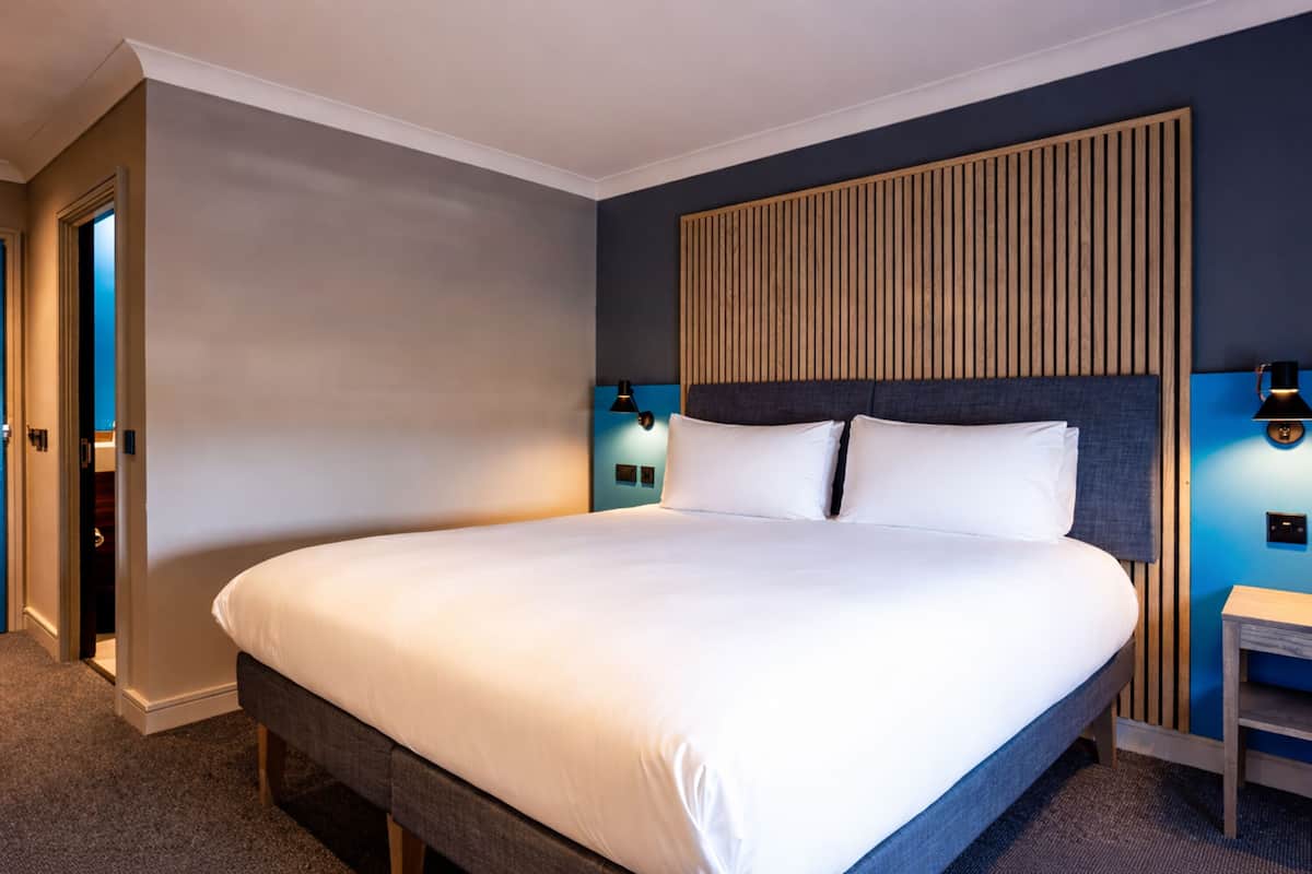 A hotel room with a large bed, a wooden headboard, two bedside lamps, and a small nightstand.