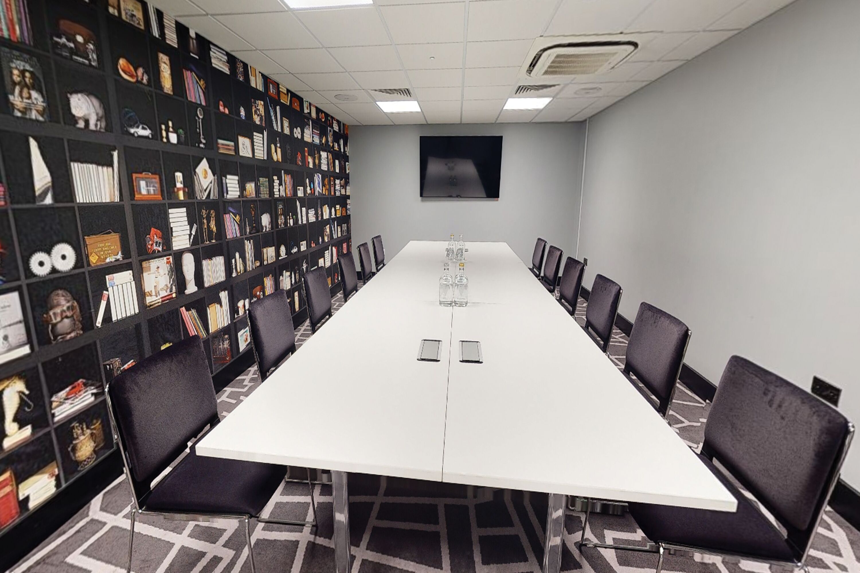 A conference room with a long table, black chairs, and a bookshelf filled with various items.