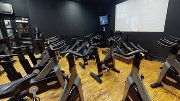 Un gimnasio con varias bicicletas estáticas, una pantalla de proyección mostrando información y piso de madera.