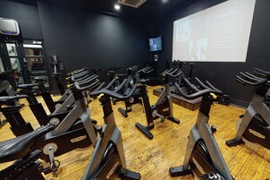 Un gimnasio con varias bicicletas estáticas, una pantalla de proyección mostrando información y piso de madera.
