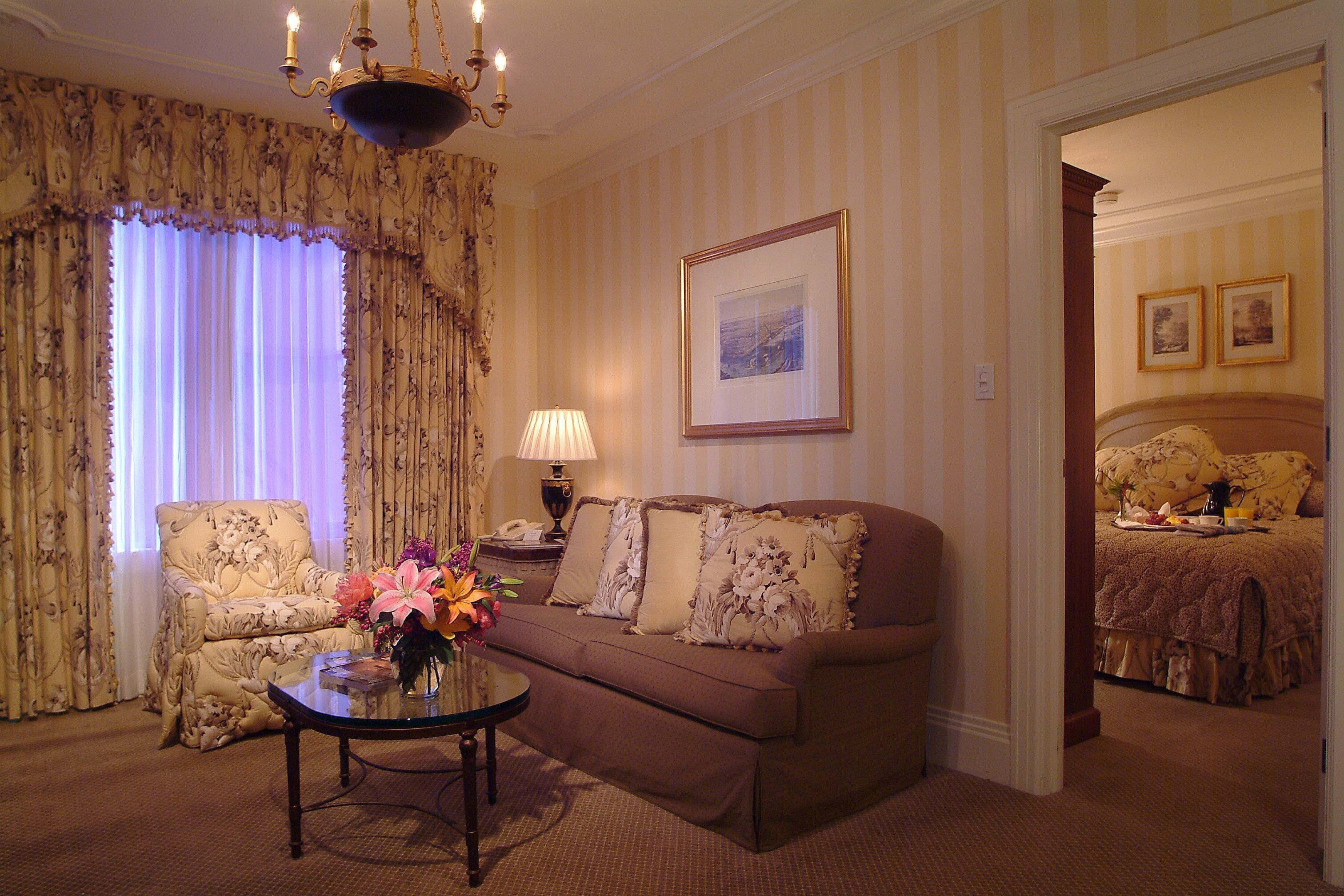 Une pièce avec un canapé, un fauteuil et une table sur laquelle est posé un vase de fleurs. On y voit également une lampe, un tableau encadré au mur et, au fond, un lit.