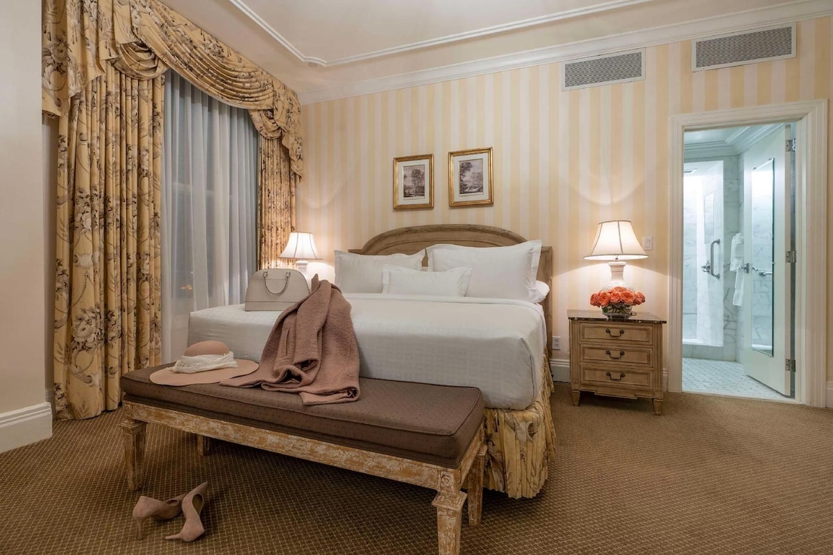 A hotel room with a bed, bedside tables, a bench, and a bathroom visible through an open door.