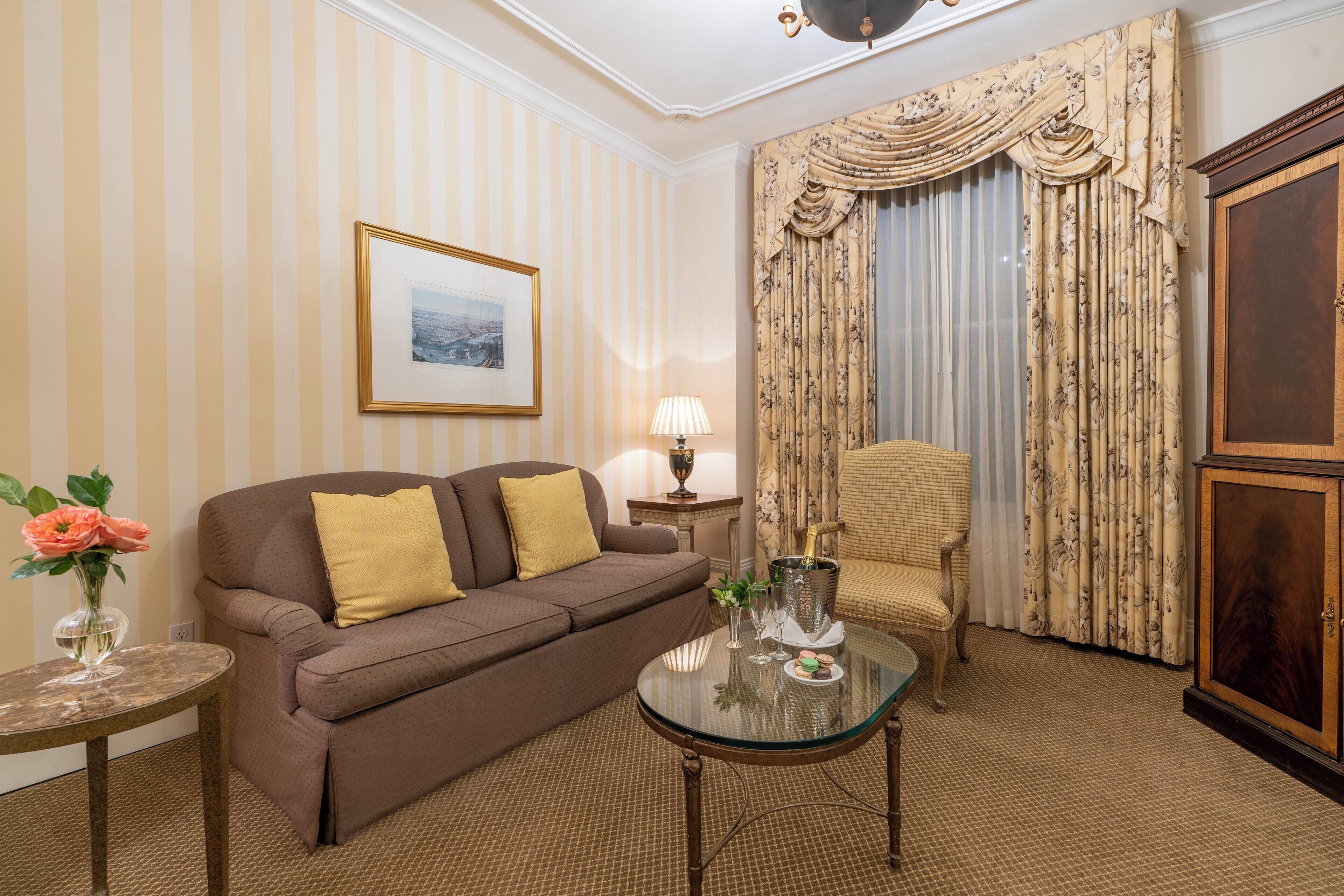 A room with a striped wallpaper, a brown sofa with yellow cushions, a glass coffee table with a bottle and glasses, a side table with a lamp, and a chair with a floral curtain.