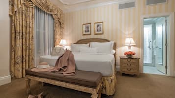 A hotel room with a large bed, a bench, a nightstand with a lamp, and a door leading to a bathroom.