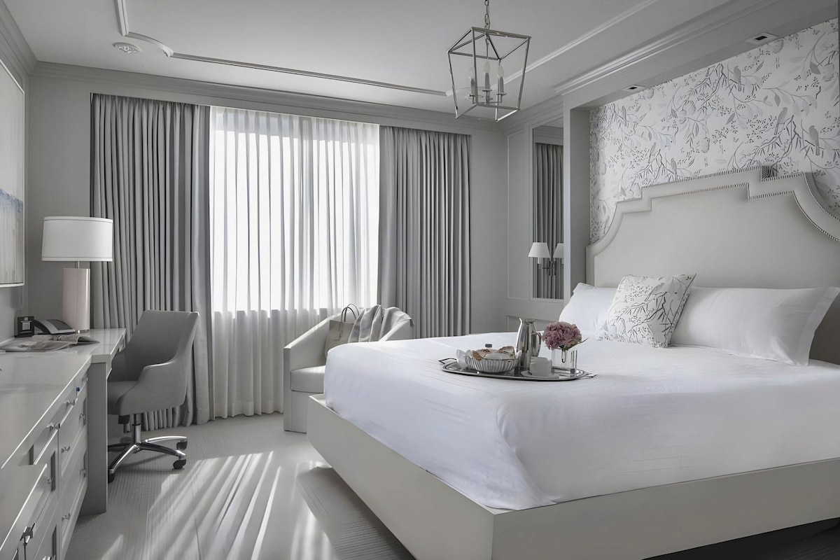 A modern hotel room with a large bed, a desk, a chair, and a nightstand.
