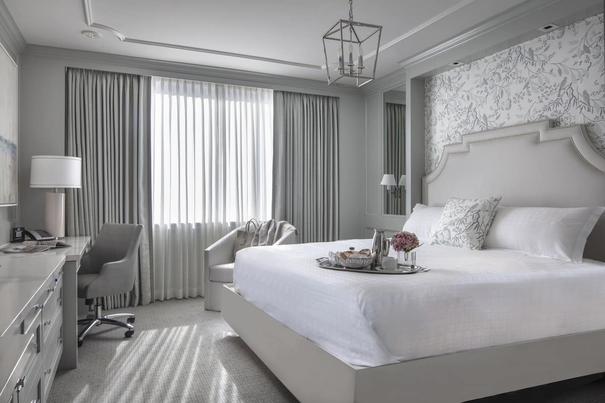 A modern hotel room with a large bed, a desk, a chair, and a desk lamp.
