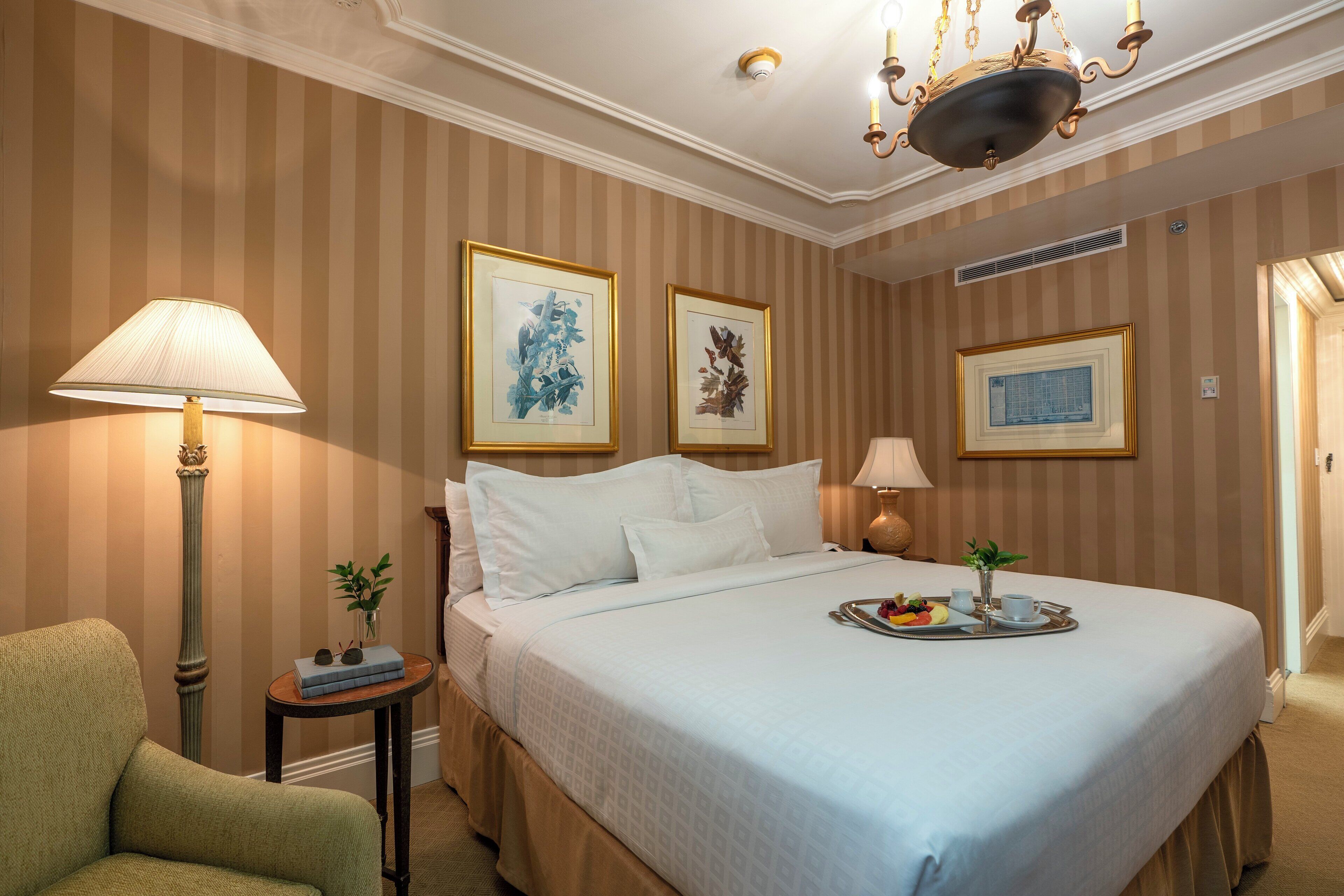 Una habitación de hotel con una cama grande, una silla, una lámpara y cuadros en la pared.