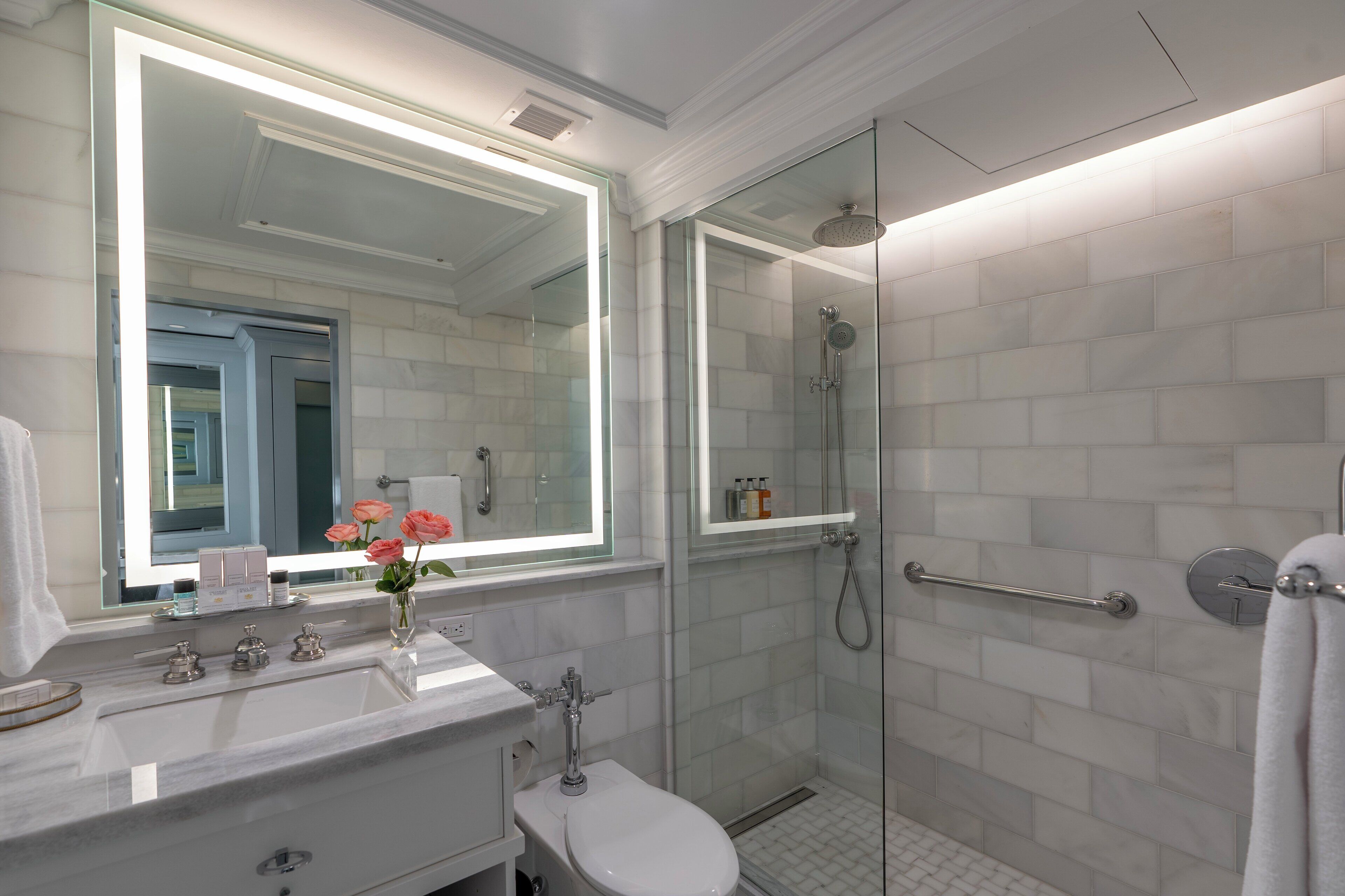 A modern bathroom with a large mirror, white tiles, a glass shower enclosure, and a white sink.