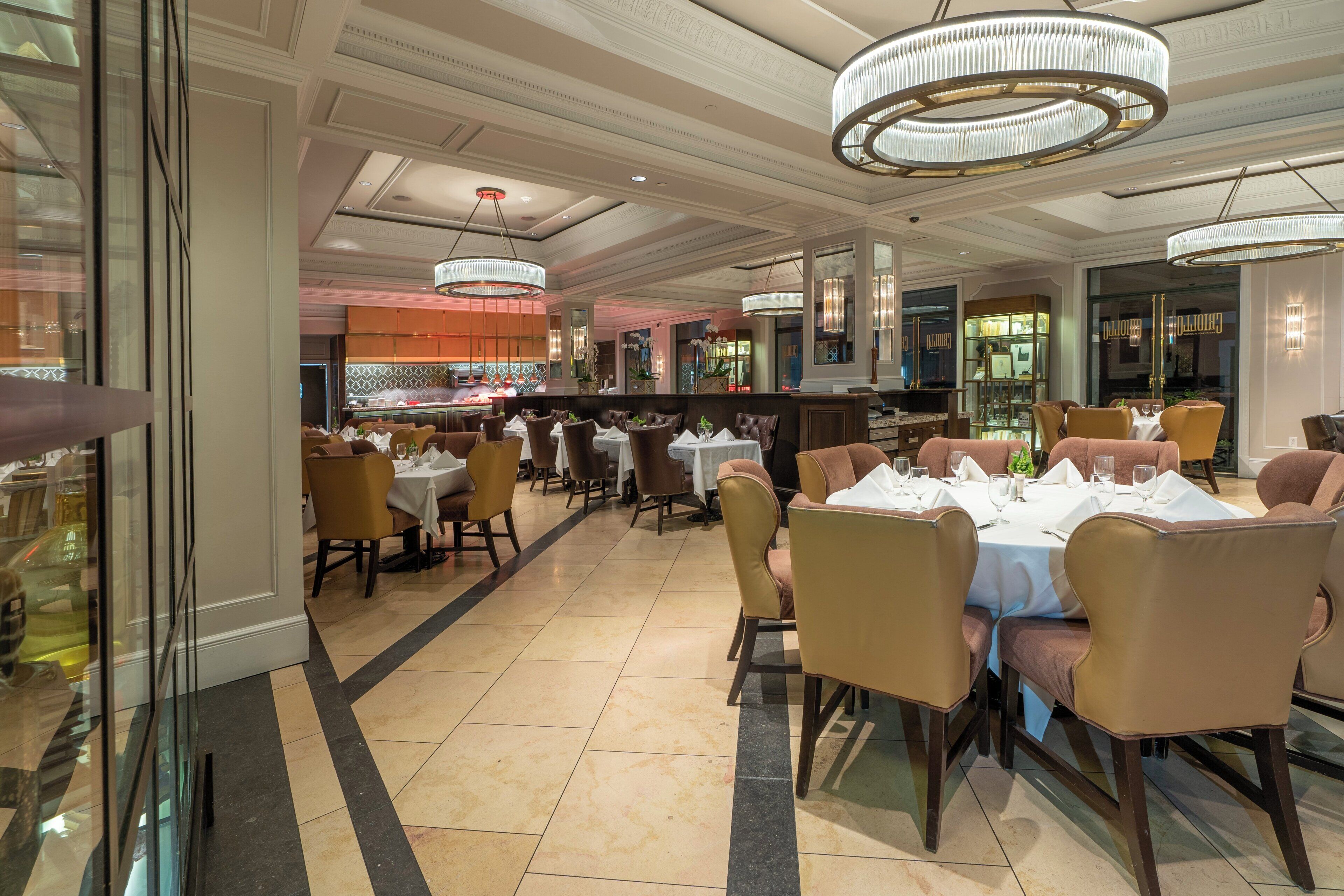 A spacious restaurant with high ceilings and modern chandeliers. Tables are set with white tablecloths and multiple chairs.