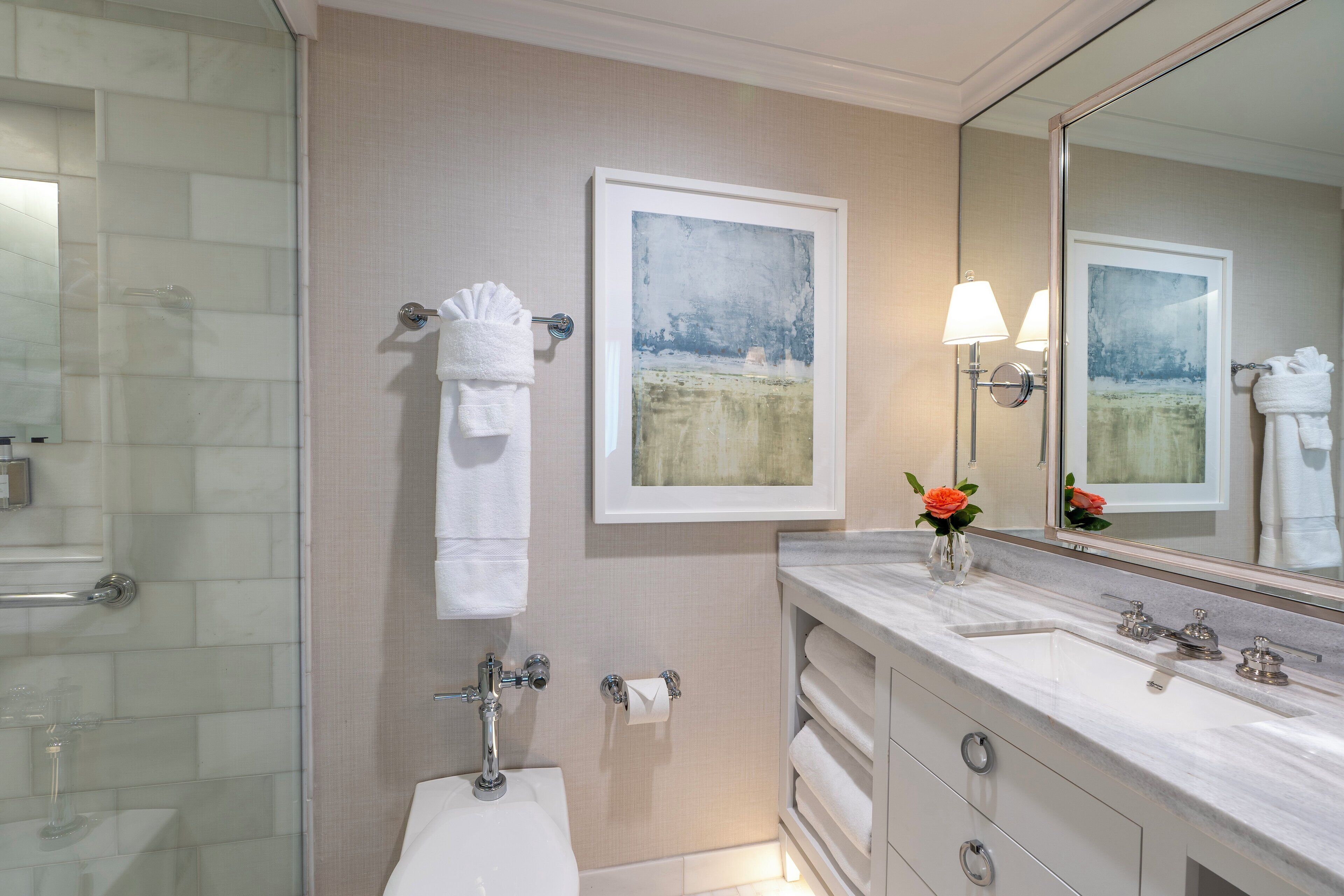 Un baño moderno con cabina de ducha de vidrio, un lavamanos blanco con encimera de mármol y dos cuadros de paisajes colgados sobre el espejo.