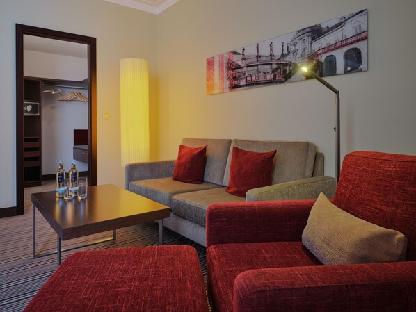 A hotel room with a grey sofa, red armchairs, a wooden coffee table, and a painting of a building on the wall.