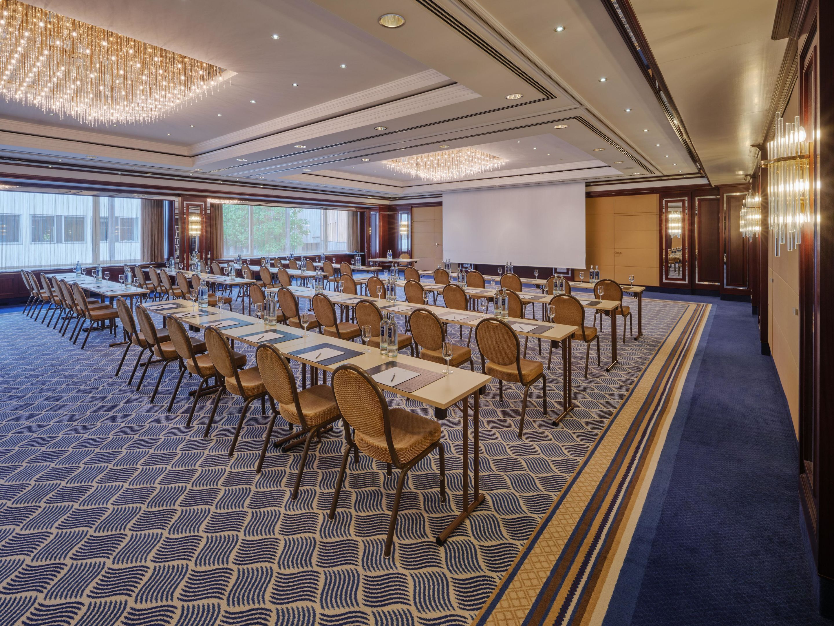 A conference room with rows of tables and chairs, large windows, and a chandelier.