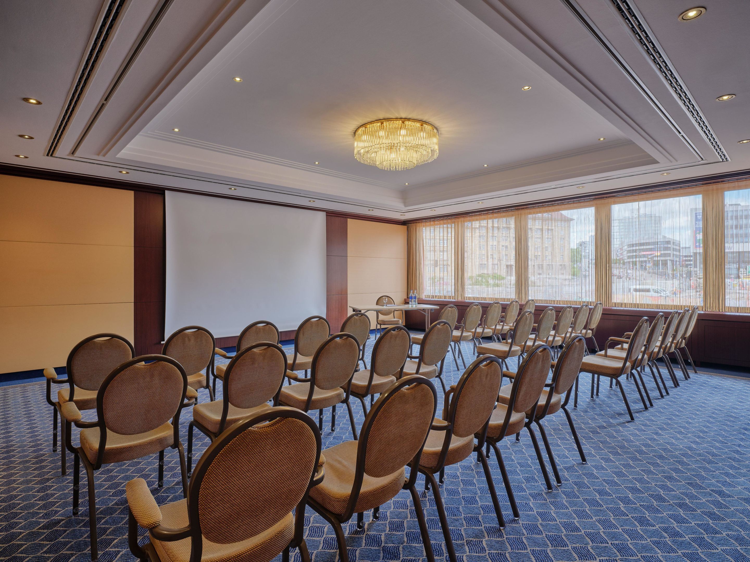 A conference room with rows of chairs, a projector screen, and a large window with a city view.