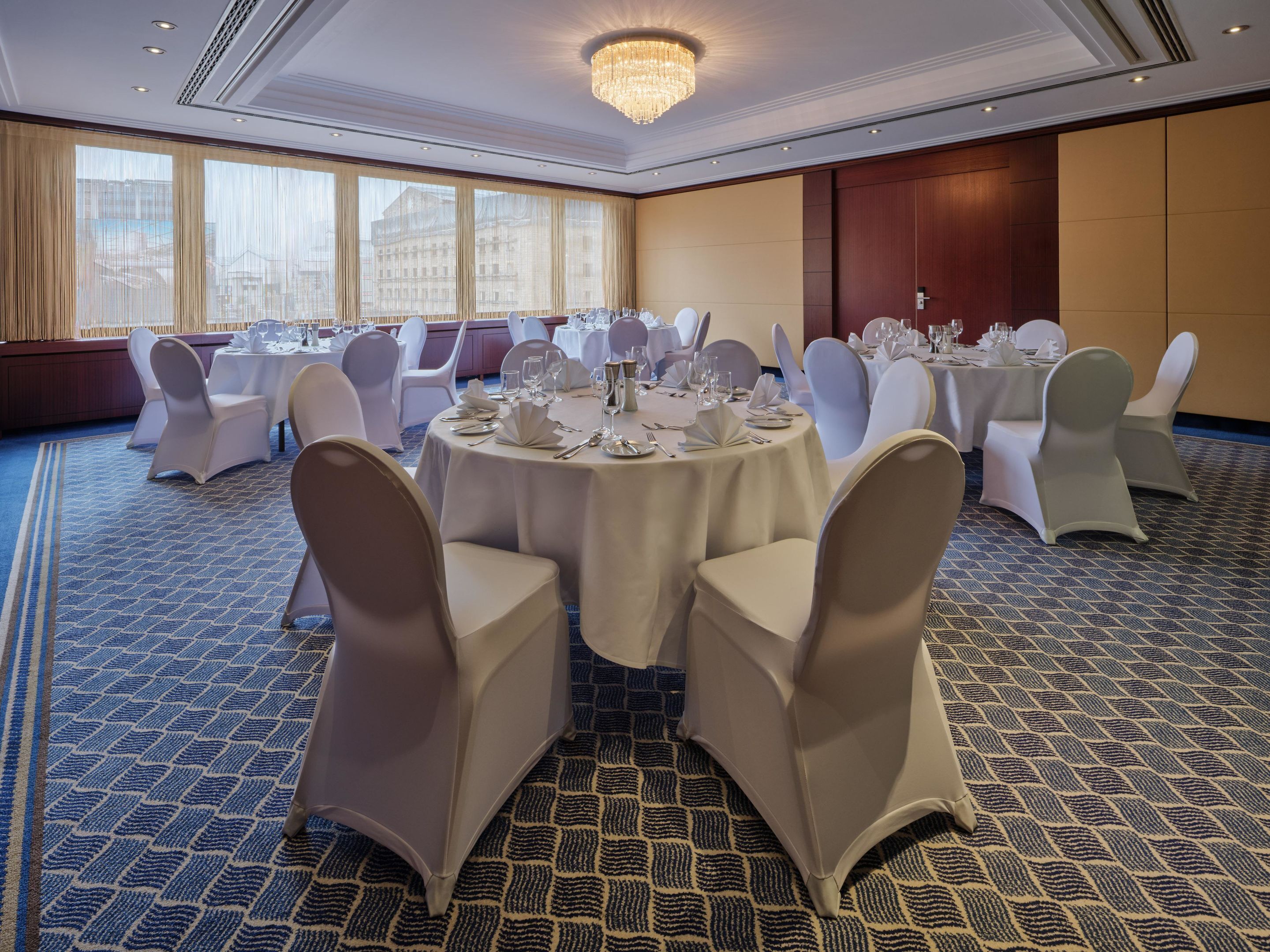 Un salón de banquetes con mesas redondas dispuestas para comer, sillas blancas y una araña.