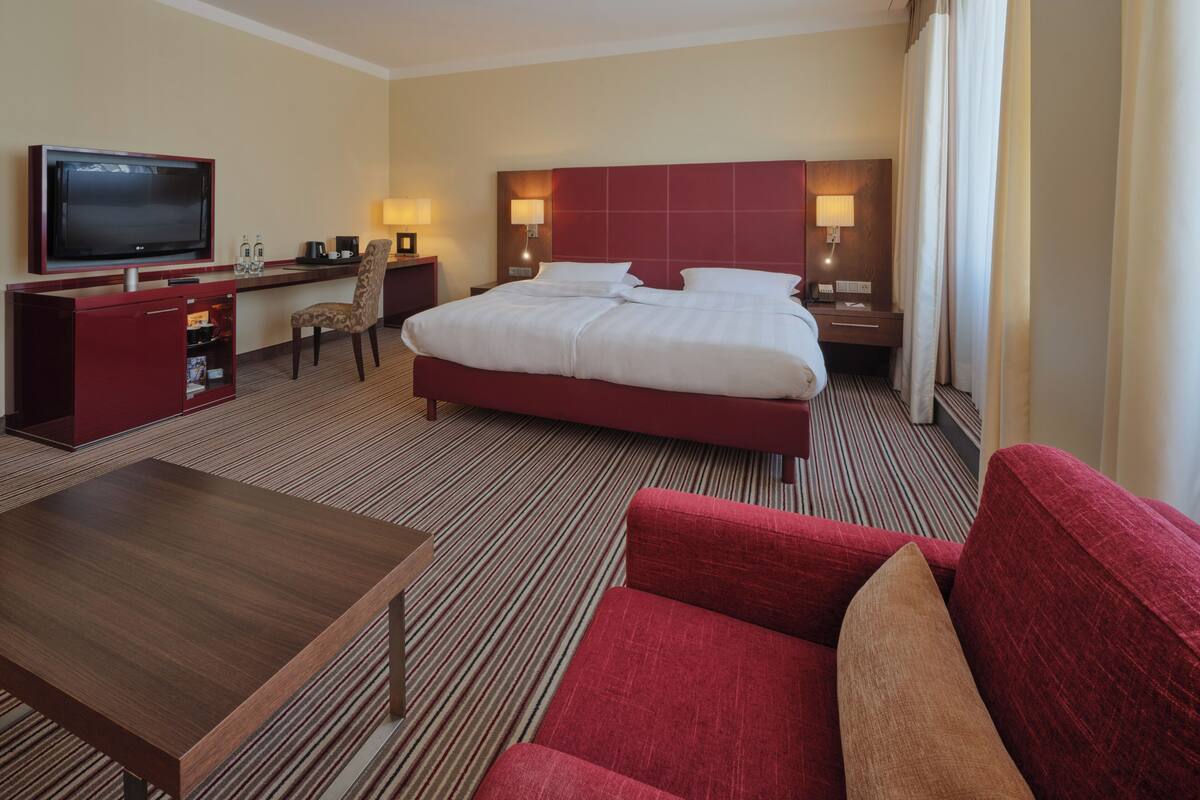 Habitación de hotel con una cama grande, un escritorio con televisor, una silla y un sofá rojo.
