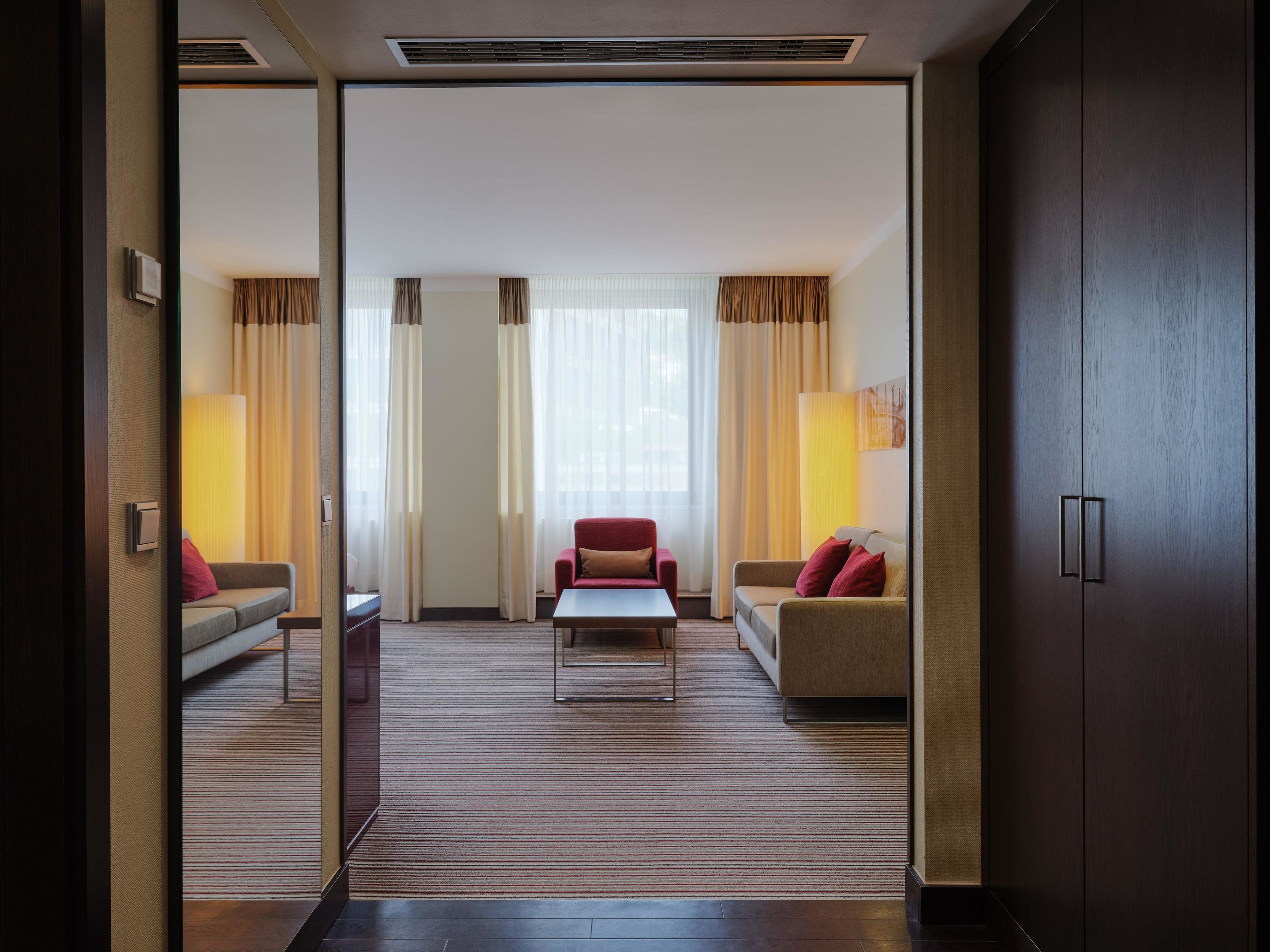 A hotel room with a sofa, a red armchair, a coffee table, and a large window with curtains.