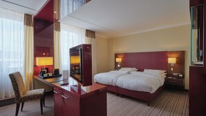 A hotel room with a large bed, a desk with a chair, a TV, and a minibar. - Steigenberger Graf Zeppelin (Stuttgart)