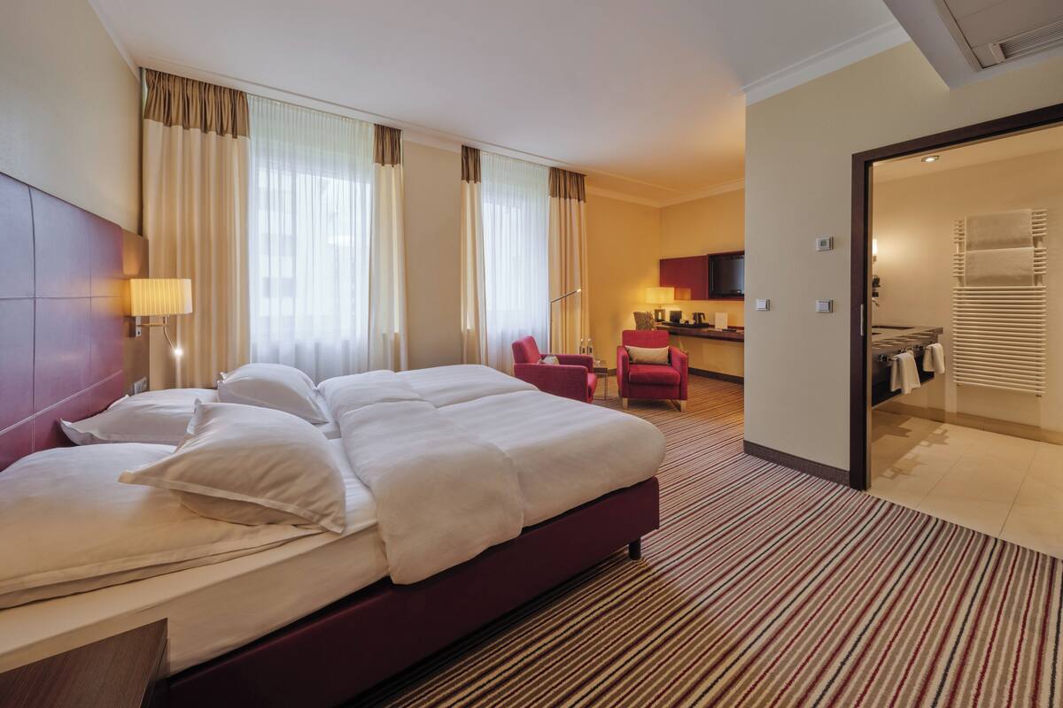 Habitación de hotel con una cama grande, un escritorio, una silla y un baño visible a través de una puerta abierta.