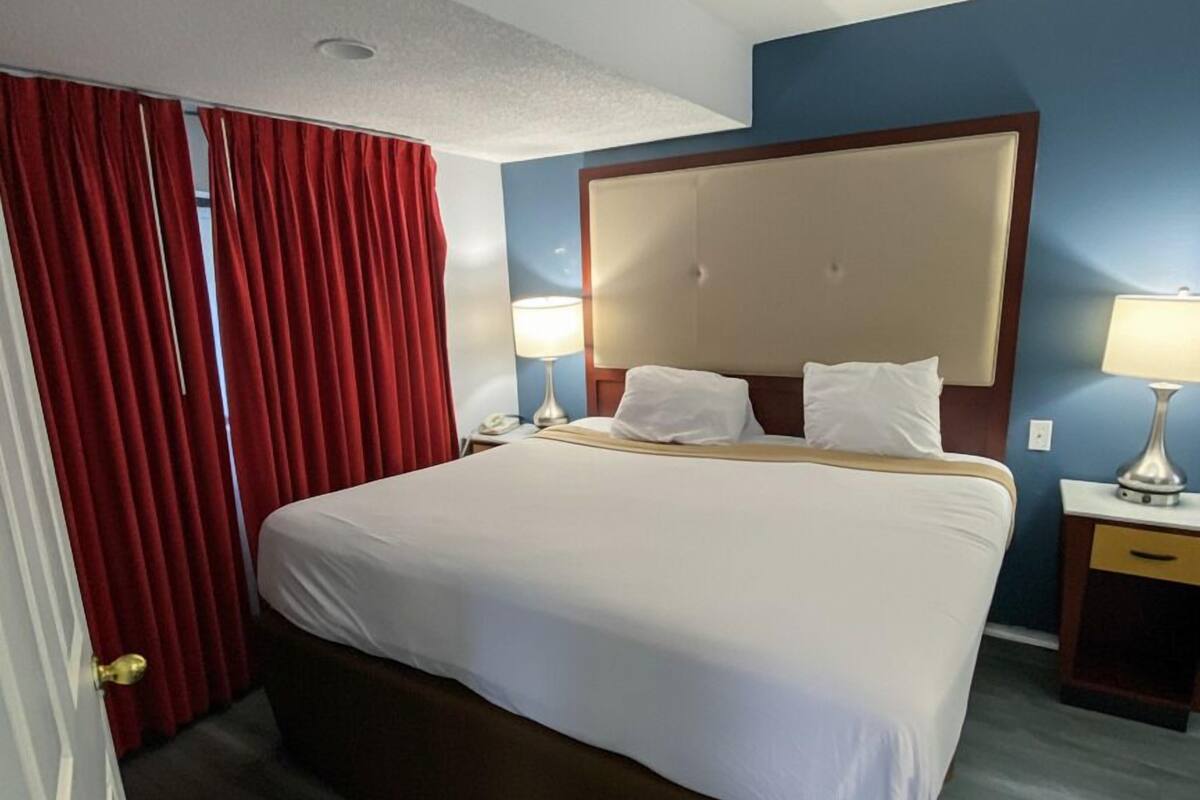 A hotel room with a large bed, red curtains, two bedside lamps, and a wooden nightstand.