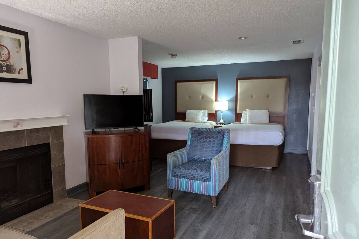 A hotel room with a television, a fireplace, a striped armchair, two beds, and a nightstand with a lamp.