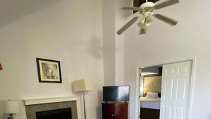 A living room with a black leather sofa, a fireplace, a TV on a wooden stand, a ceiling fan, and a door leading to another room. - Extended Stay of Charleston (North Charleston)