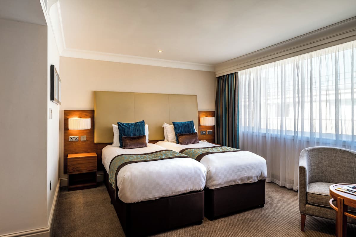 A hotel room with two beds, a large headboard, bedside lamps, a chair, and a view of the outdoors through sheer curtains.