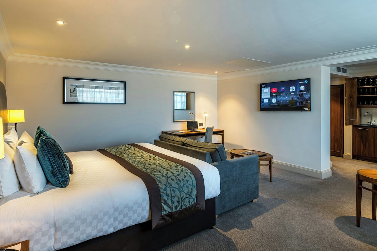 A hotel room with a large bed, a desk, a TV, and a kitchenette.