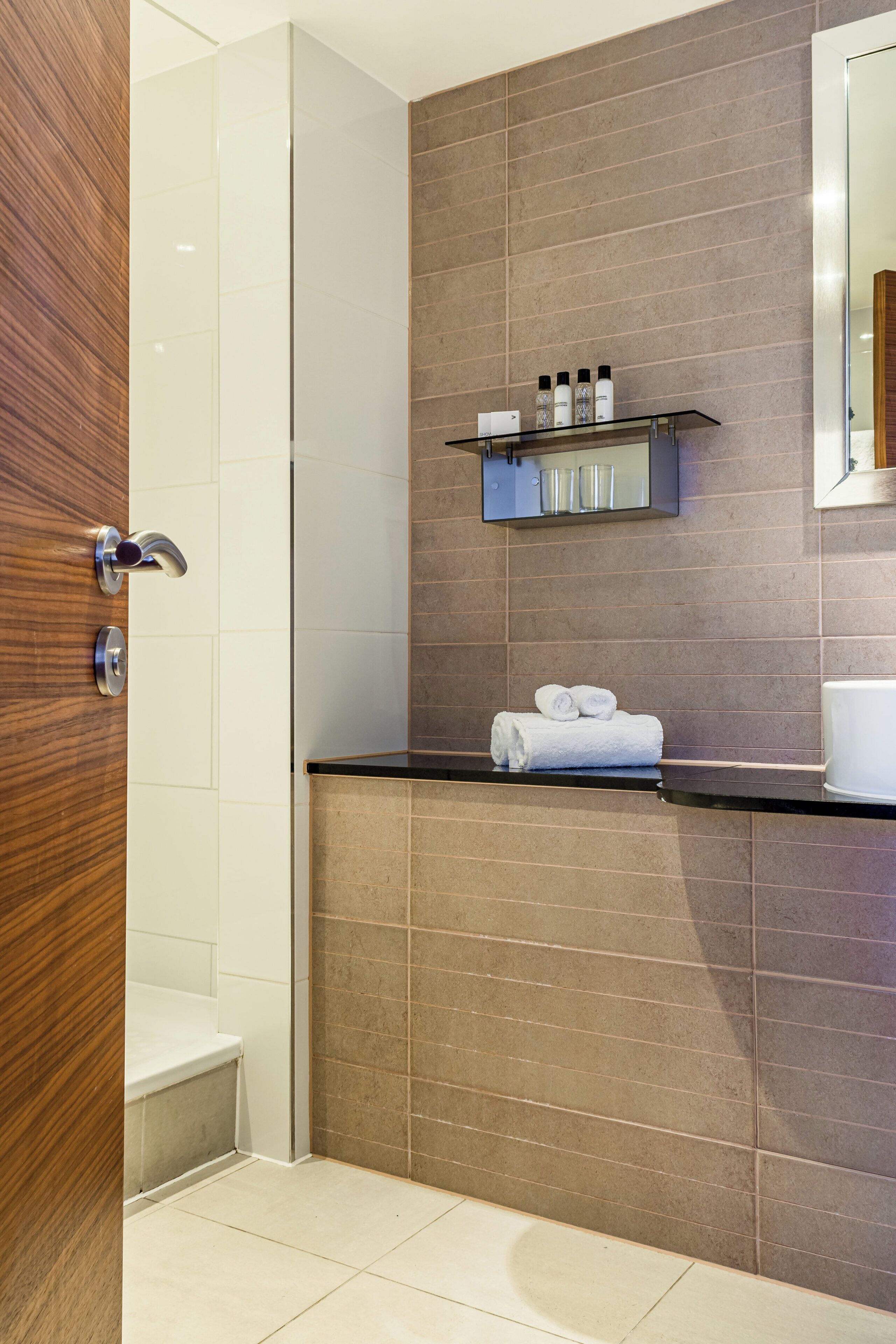 Un baño moderno con puerta de madera, cabina de ducha de vidrio, encimera negra y espejo sobre ella.