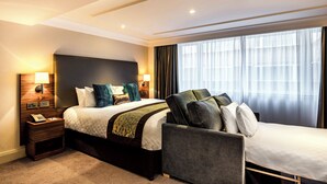 Habitación de hotel con una cama extragrande, un sofá cama independiente, una mesita de noche con lámpara y un ventanal con cortinas.