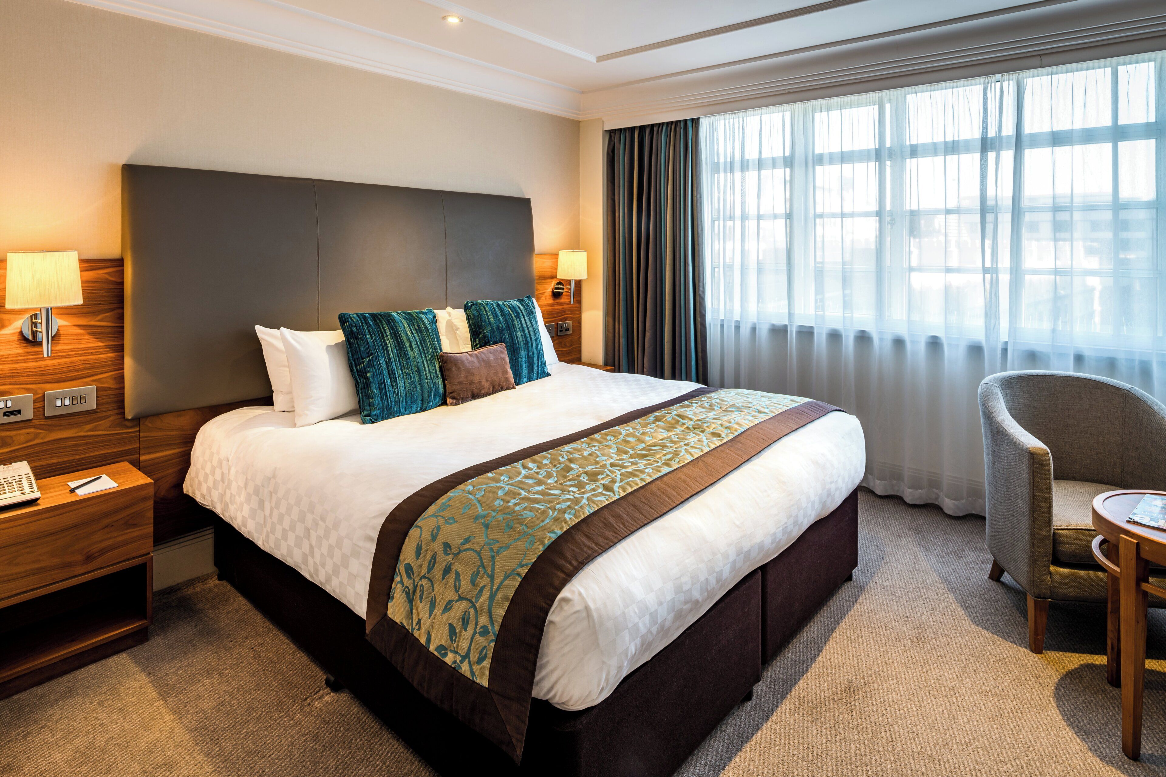 A hotel room with a large bed, two bedside tables with lamps, a chair, and a window with curtains.
