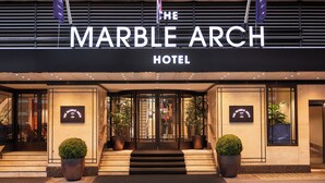 Entrada del Marble Arch Hotel con fachada negra, amplias puertas de cristal y plantas en macetas.