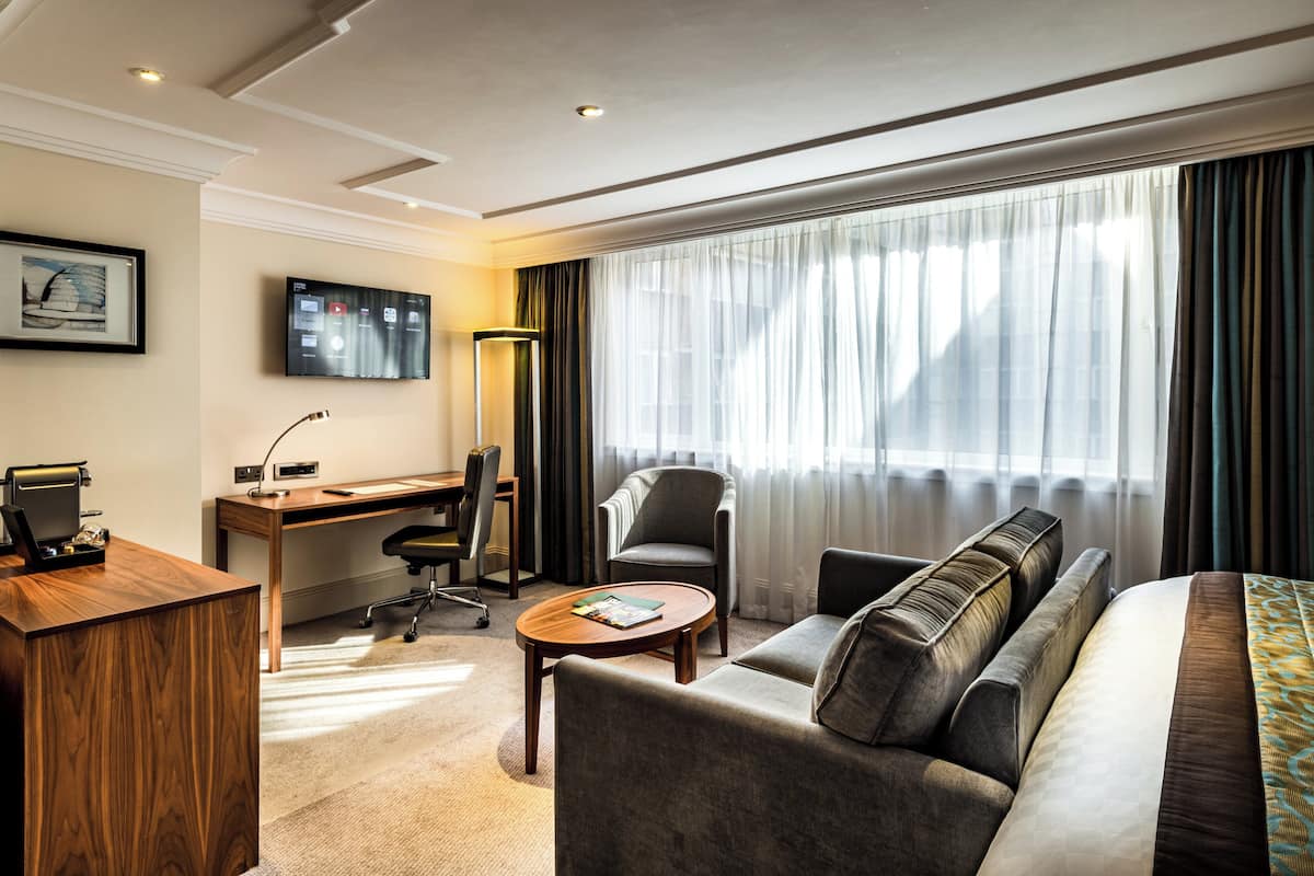A hotel room with a wooden desk, a comfortable sofa, a small round table, and a TV mounted on the wall.
