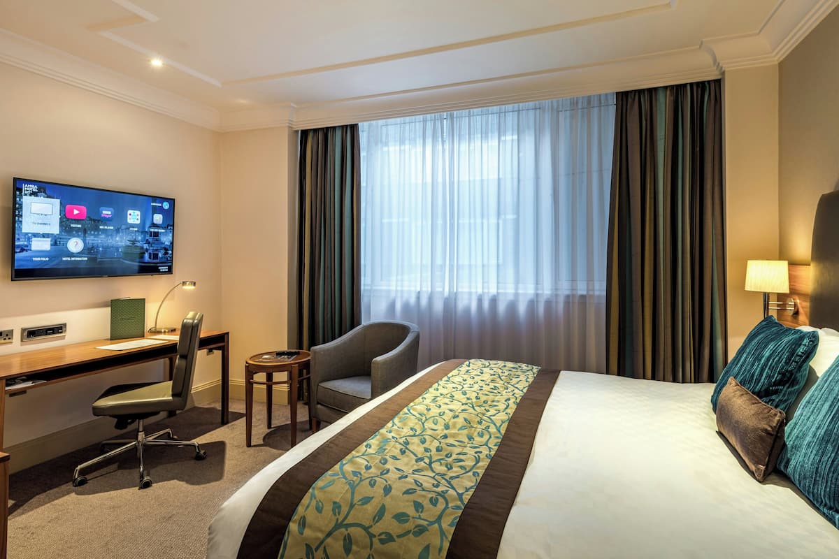 A hotel room with a bed, a desk with a chair, a television, and a window with curtains.