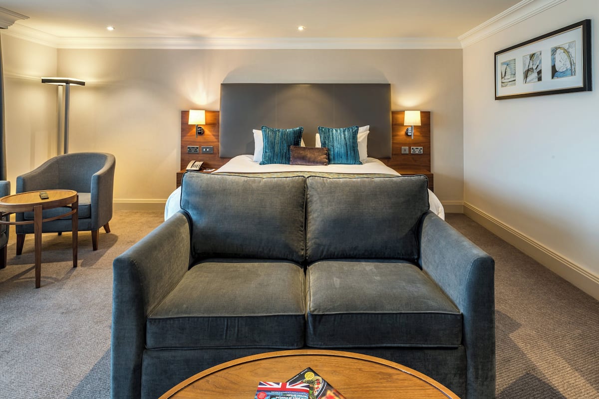 A hotel room with a large bed, two bedside tables with lamps, a grey sofa, a round coffee table, and a chair.