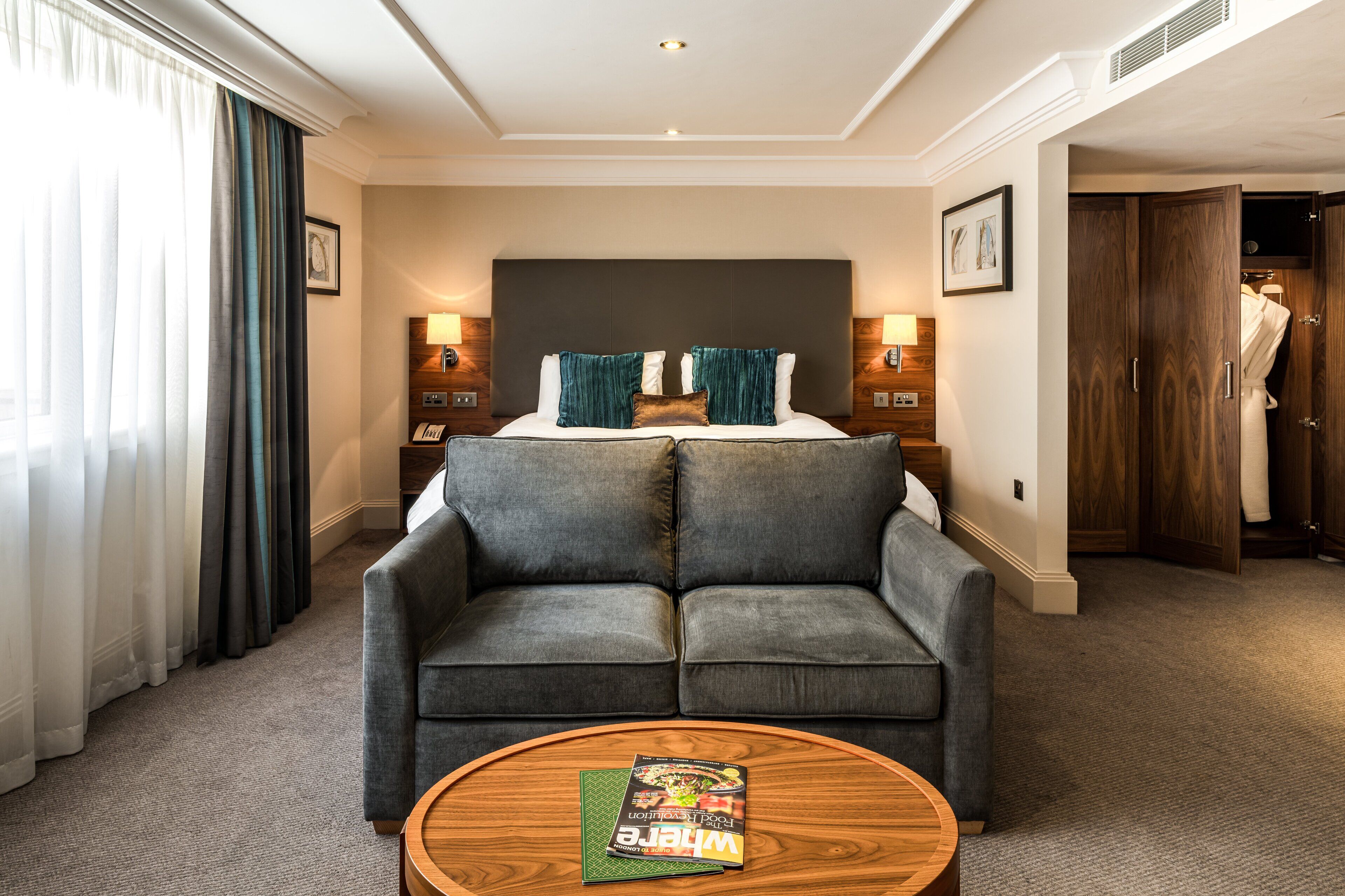 Une chambre d’hôtel avec un grand lit, deux tables de chevet avec des lampes, un canapé gris, une table basse en bois avec un magazine et une armoire où est suspendu un peignoir.