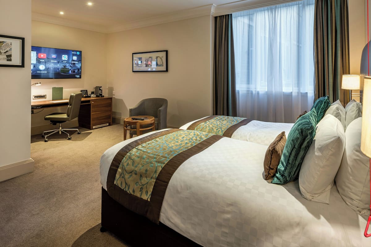 A hotel room with two beds, a desk, a chair, a TV, and a window with curtains.