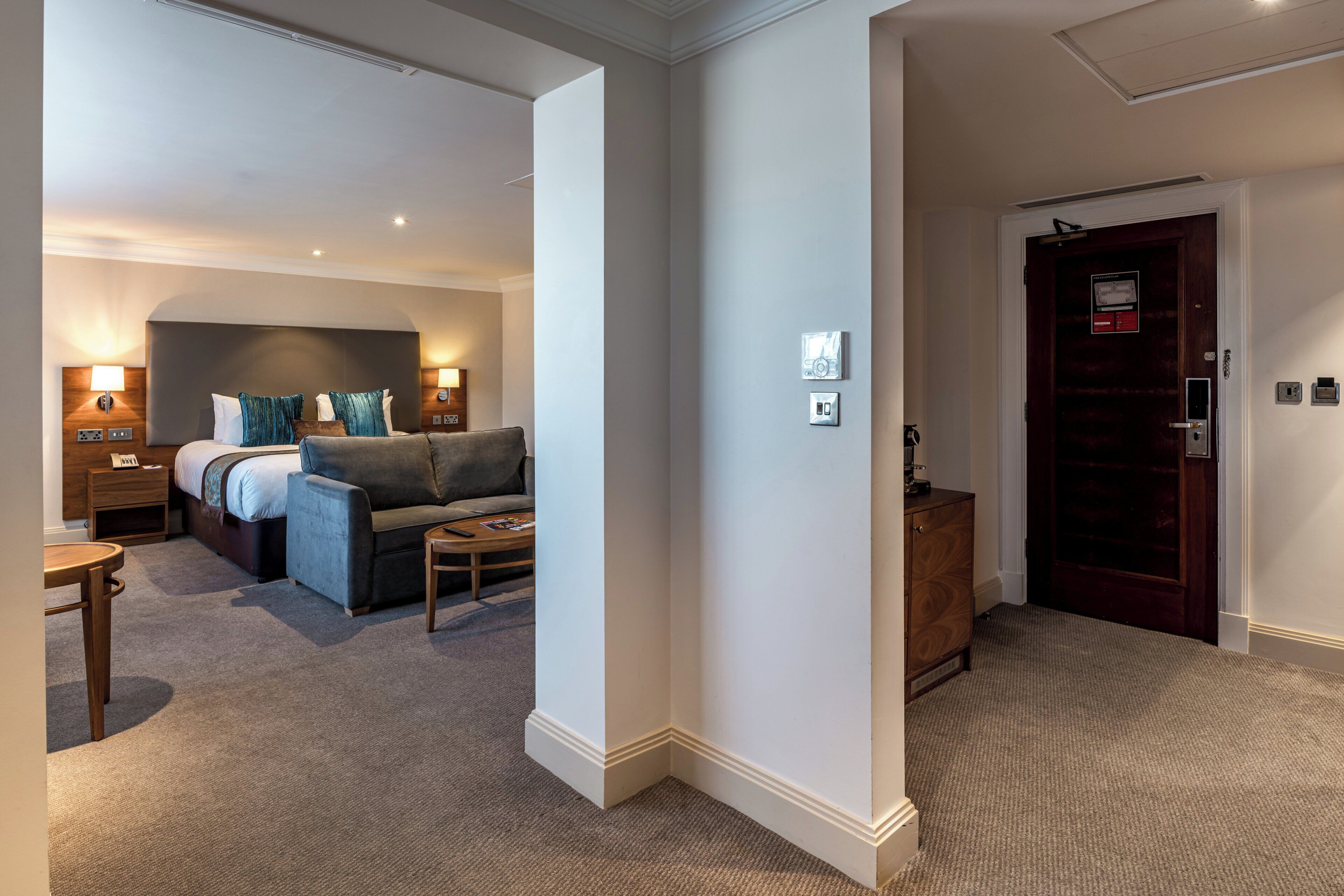 A hotel room with a bed, a sofa, a small table, and a wooden cabinet. There are wall-mounted lights and a door with a lock.