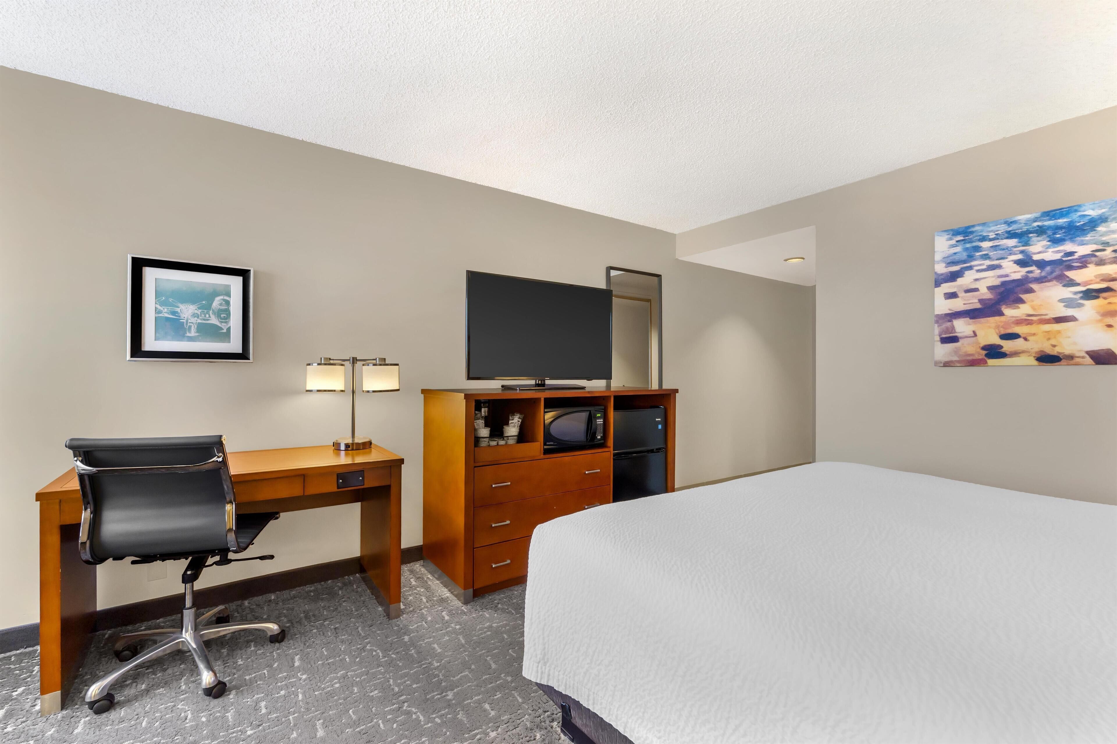 A hotel room with a bed, a desk with a chair, a dresser, a flat-screen TV, and a framed picture on the wall.