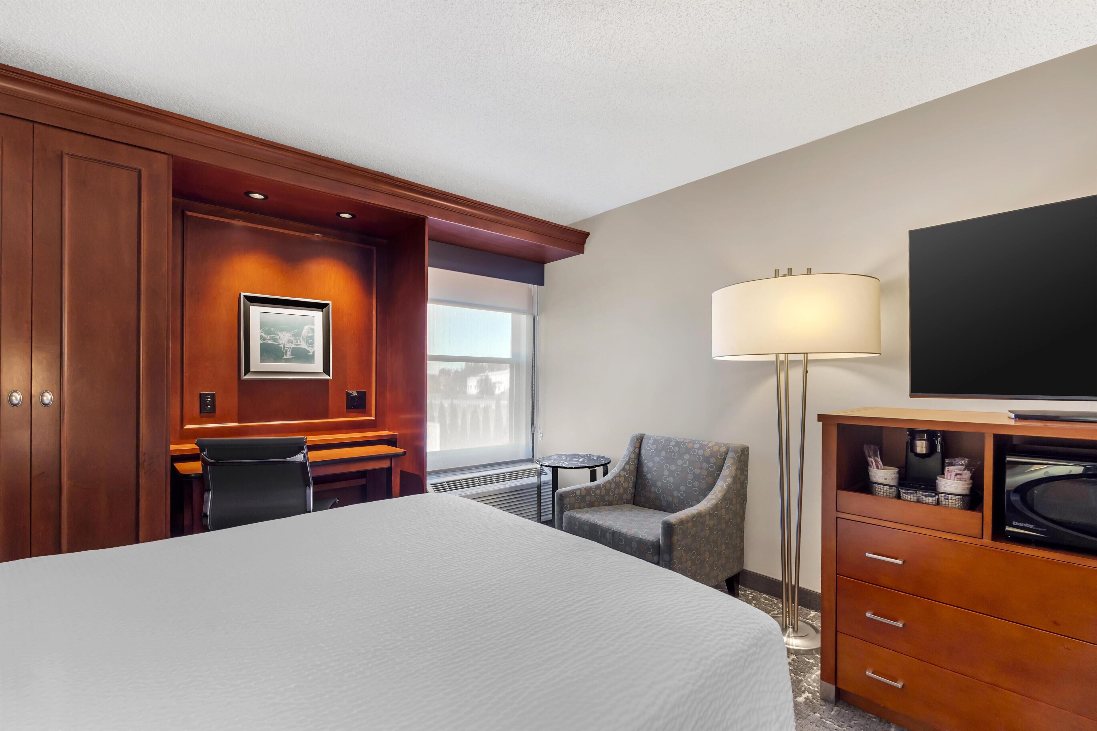 A hotel room with a large bed, a desk, a chair, a TV, a lamp, and a coffee machine.