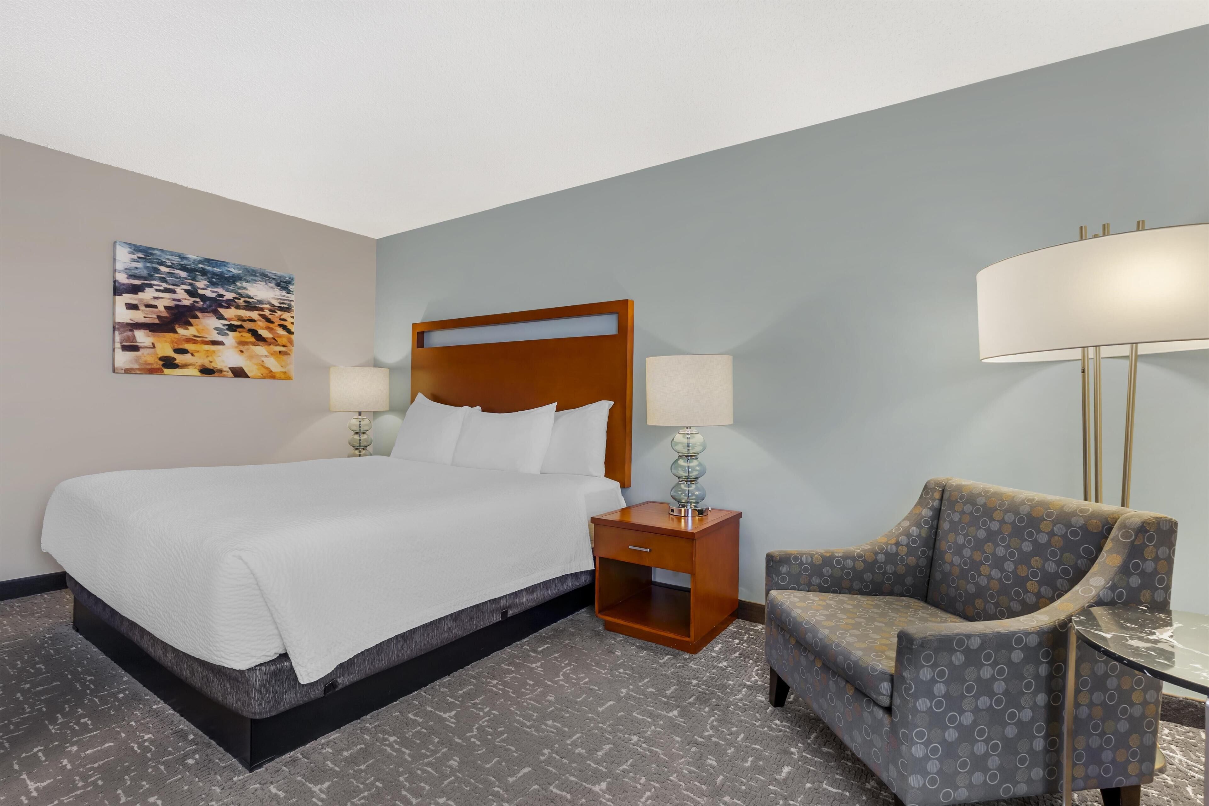 Habitación de hotel con una cama grande, una mesita de noche de madera con lámpara, un sillón estampado y una pequeña mesa.
