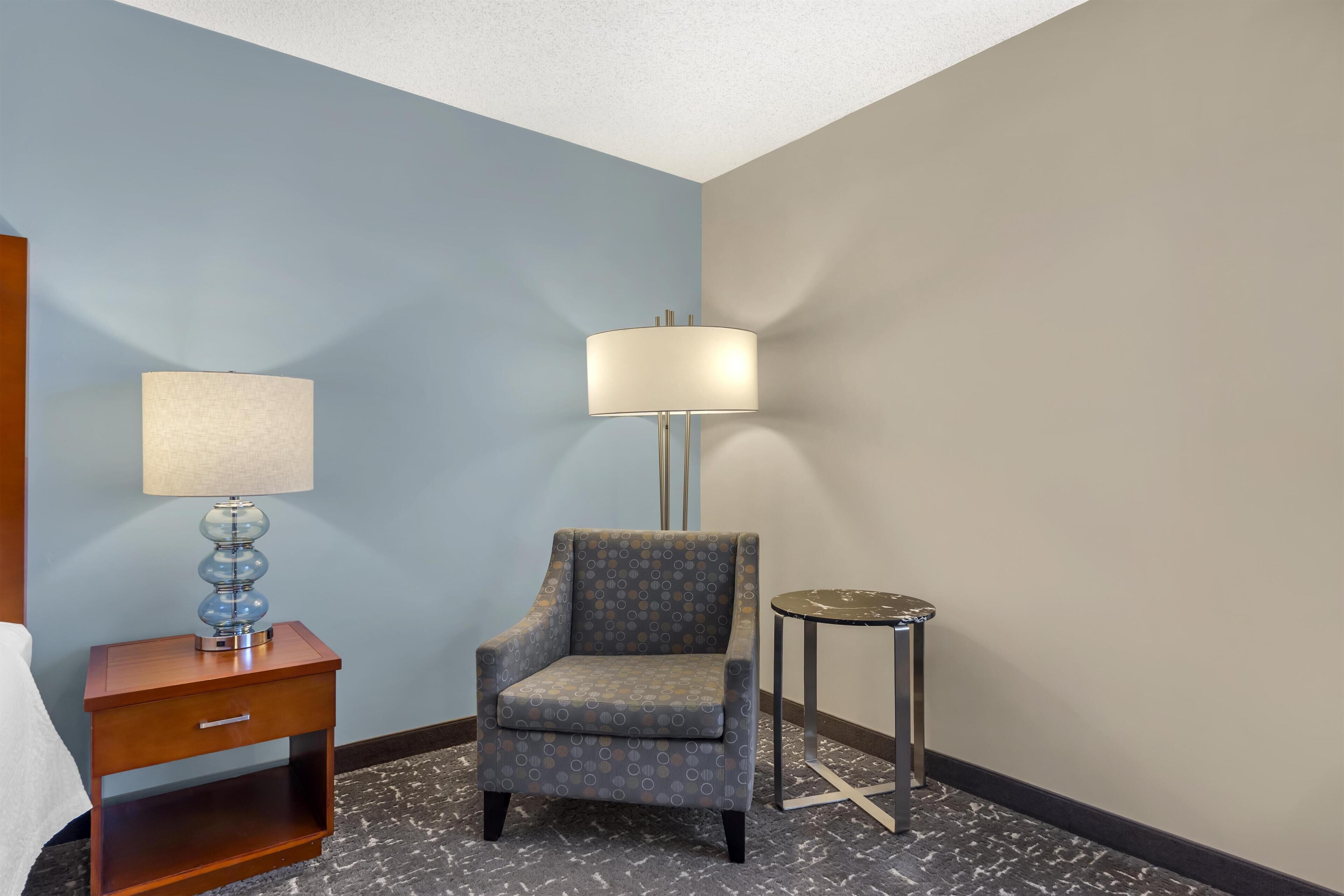A hotel room with a single armchair, a small table with a lamp, and a nightstand with a lamp.