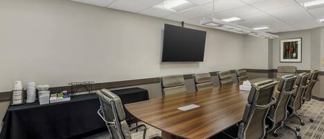 A conference room with a large wooden table, multiple chairs, a projector screen, and a table with supplies.