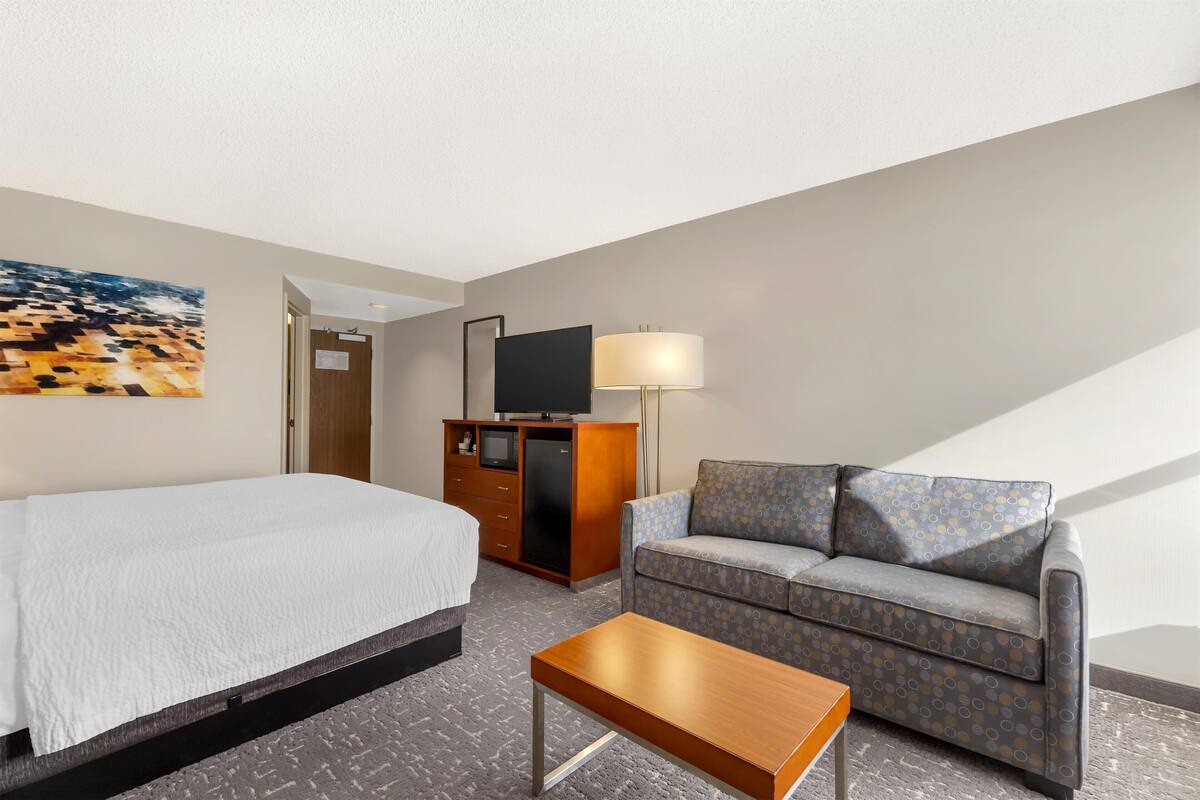 A hotel room with a bed, a sofa, a TV on a wooden cabinet, a lamp, and a painting on the wall.