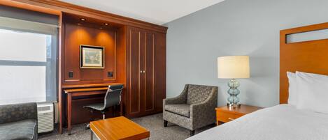 A hotel room with a bed, a desk, a chair, a small table, a lamp, and a picture on the wall.