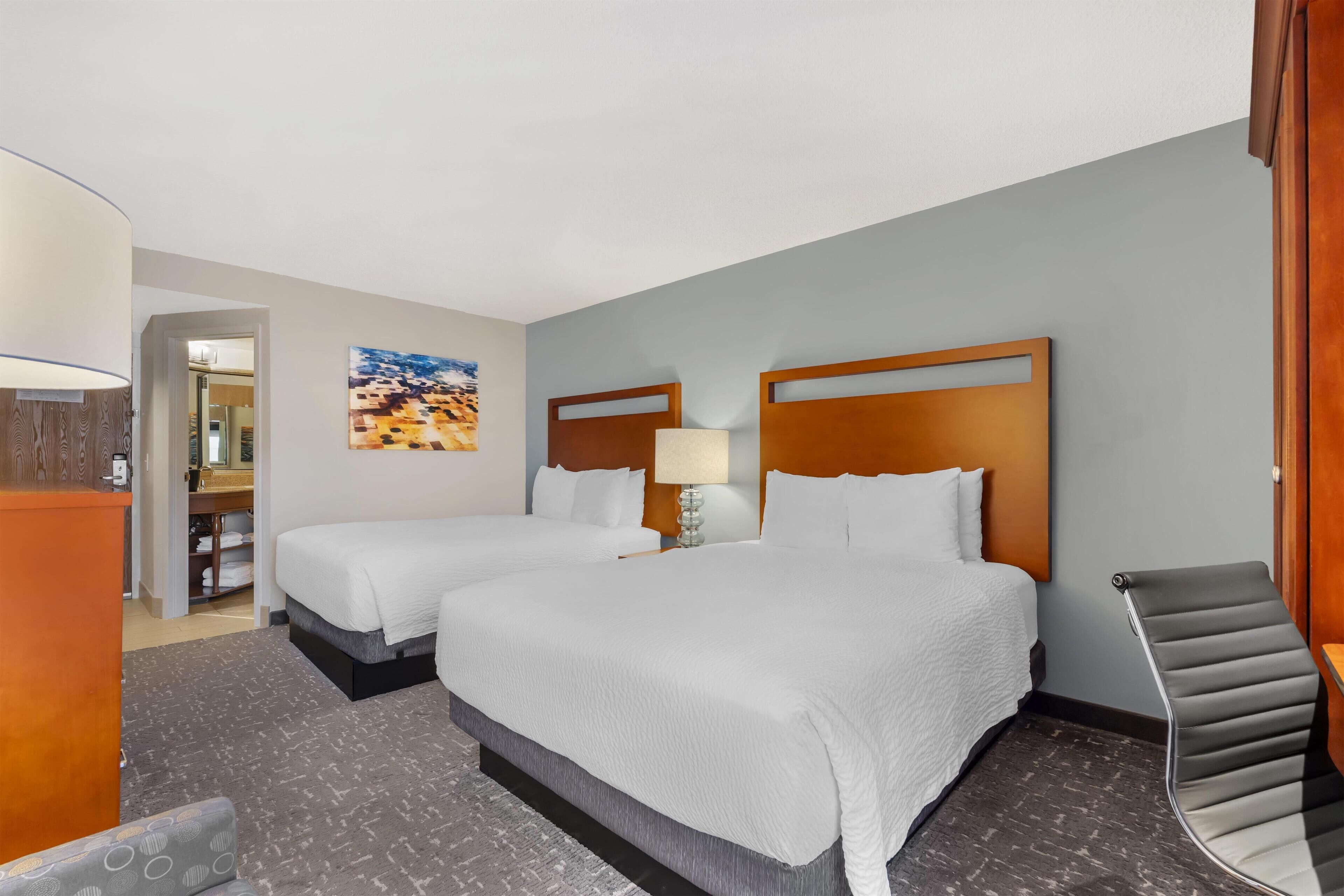 A hotel room with two beds, a desk, a chair, and a painting on the wall.