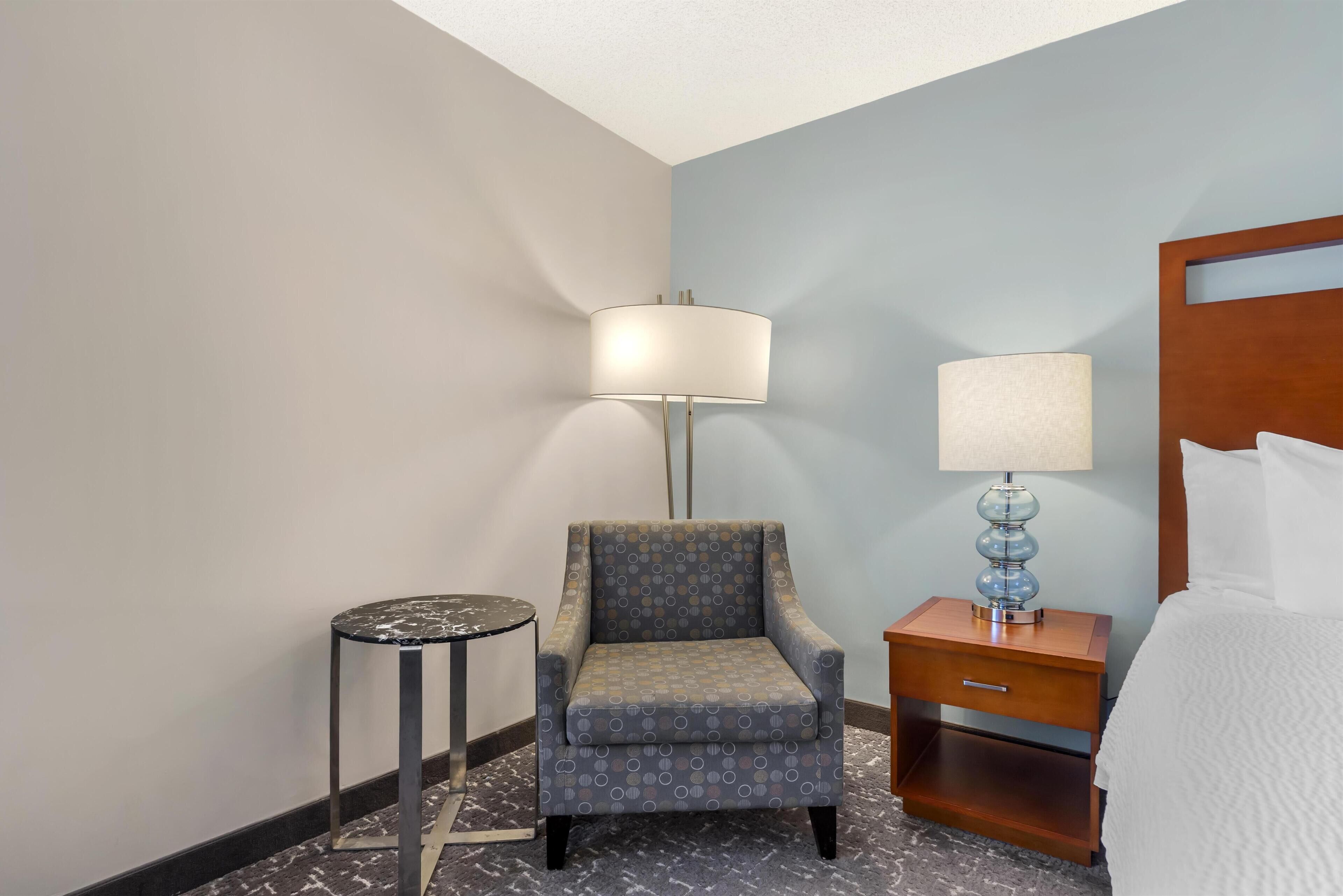 A hotel room with a grey armchair, a small round table, a bedside table with a lamp, and a bed with white bedding.